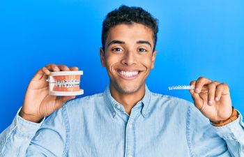 Young Afro-American man with a perfect smile holding a dental model with orthodontic braces in one hand and a clear orthodontic aligner in the other one.